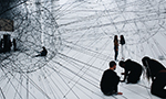 Photograph of a work of art composed of two globe-shaped structures made and lifted by steel cables. In the foreground, three people are kneeling facing where one of the cables is attached to the ground to the right of one of the globes. In the background, next to the other globe, two people are standing talking.